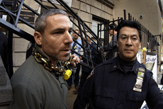 Man with bicycle chain around his neck, a steel gate behind him, a police officer to the right, looking at the camera.